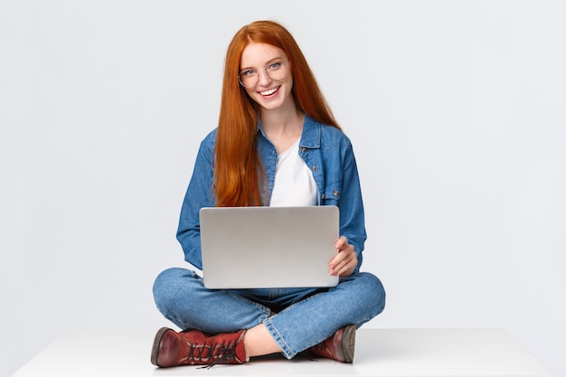 Une étudiante rousse joyeuse et impertinente, une collégienne à lunettes, assise sur les jambes croisées avec un ordinateur portable, travaillant en freelance, prépare un projet depuis le bureau, à distance sur fond blanc.