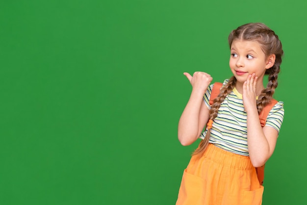 L'étudiante avec la serviette est très surprise et pointe son doigt sur le côté. une fille avec un sac sur un fond vert isolé. l'écolière montre la publicité.