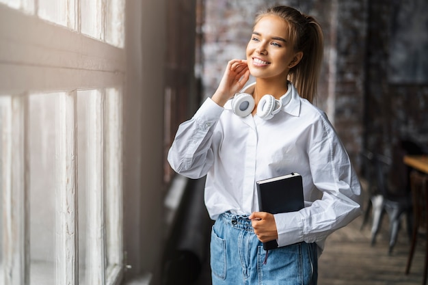 Une étudiante souriante dans une chemise blanche et des écouteurs sans fil se tient devant la fenêtre.