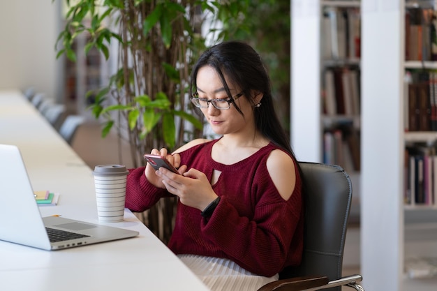 Une étudiante tergiverse dans la bibliothèque avec un chat sur smartphone dans les médias sociaux ne se prépare pas à l'examen