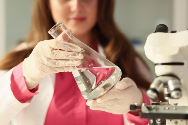 Une étudiante tient une fiole en verre avec de l'eau propre dans les mains d'une jeune femme concentrée dans des gants en caoutchouc