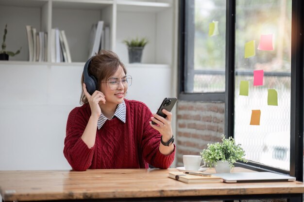 Photo Étudiante universitaire asiatique utilisant un ordinateur portable et un téléphone avec des écouteurs tout en étudiant lire des messages et saluer des amis par appel vidéo