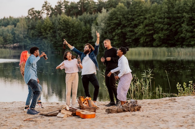 Des étudiants amis de différentes nationalités et couleurs de peau se rencontrent sur la plage au bord d'un feu de joie au bord du lac