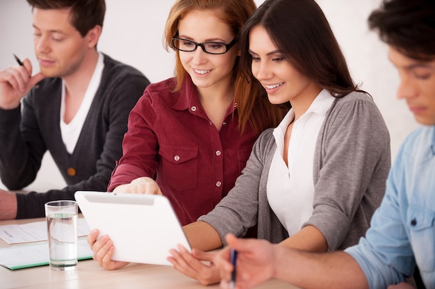 Les étudiants étudient ensemble. Groupe de jeunes assis ensemble à la table tandis que deux femmes gaies regardent la tablette numérique et sourient