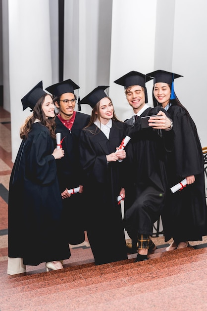 Des étudiants heureux en robes de fin d'études se font un selfie et sourient tout en tenant des diplômes.