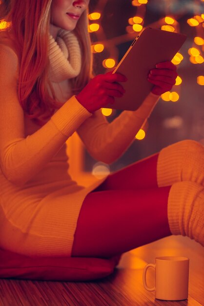Examiner sa toute nouvelle tablette. Image recadrée d'une belle jeune femme en pull blanc tenant une tablette numérique alors qu'elle était assise par terre à la maison avec des lumières de Noël défocalisées en arrière-plan