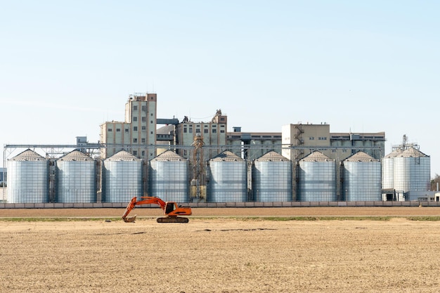 L'excavatrice se dresse sur fond de silos d'argent Usine de fabrication d'agro pour le traitement, le séchage, le nettoyage et le stockage des produits agricoles Grands barils de fer Ascenseur de grenier