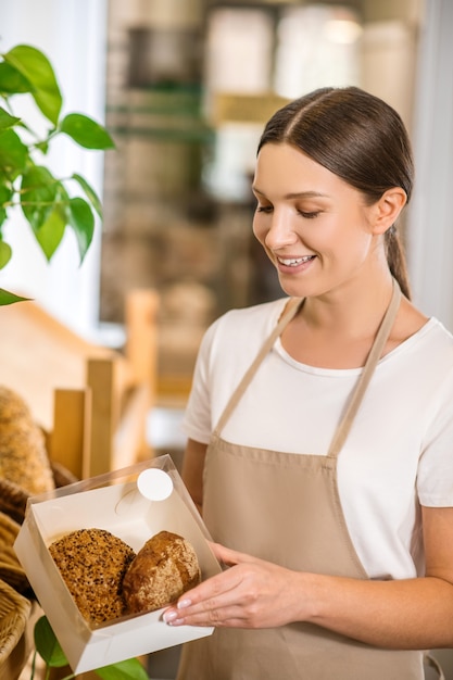 Excellent résultat. Jolie femme intéressée en tshirt et tablier à la recherche avec bonheur de la boîte avec des petits pains dans ses mains en mini boulangerie