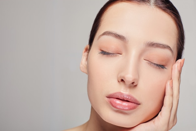 Une excellente fille sophistiquée avec des cheveux noirs et une peau propre et délicate rayonnante sur le gris. Lady a mis sa tête sur sa main. Regarde droit. Cosmétologie, soin du visage.