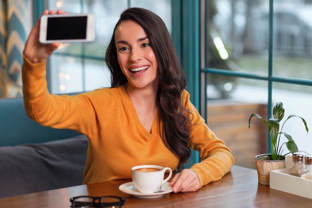 Excellente posture. Très heureuse femme merveilleuse qui monte le téléphone pour prendre selfie et boire du café