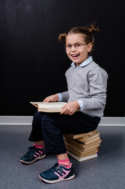 Excité adorable écolière à lunettes et vêtements décontractés assis sur une pile de livres tout en lisant l'un d'eux devant la caméra contre le tableau noir