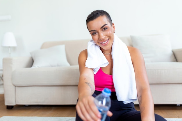 Excité athlétique femme noire en vêtements de sport riant assis sur un tapis de yoga au sol tenant une bouteille d'eau avec une serviette sur ses épaules