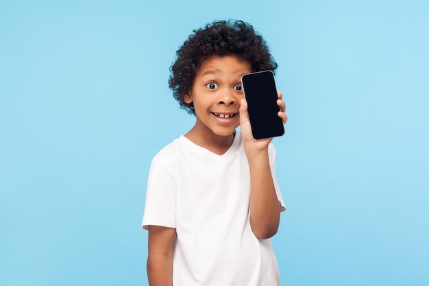 Excité drôle adorable petit garçon aux cheveux bouclés tenant un téléphone portable près du visage, enfant satisfait se réjouissant du nouveau téléphone portable et regardant avec un sourire heureux. studio d'intérieur tourné isolé sur fond bleu