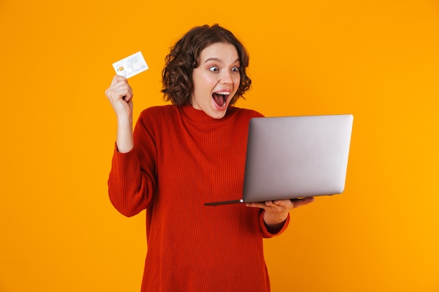 excité émotionnelle jeune jolie femme posant isolé sur mur jaune à l'aide d'un ordinateur portable tenant une carte de crédit.