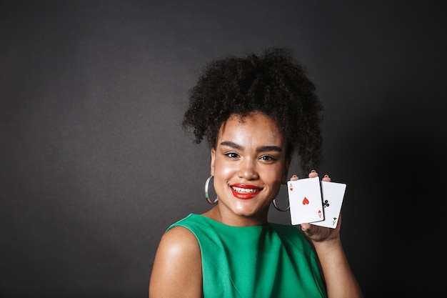 Excité femme africaine vêtue d'une robe montrant des cartes à jouer isolées sur un espace noir