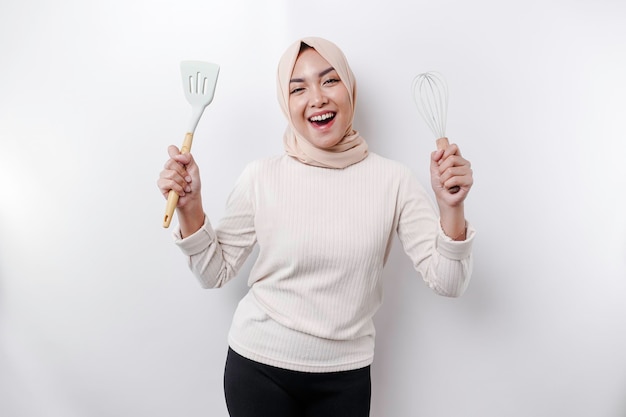 Excité femme musulmane asiatique portant un foulard tenant des ustensiles de cuisine et souriant isolé sur fond blanc
