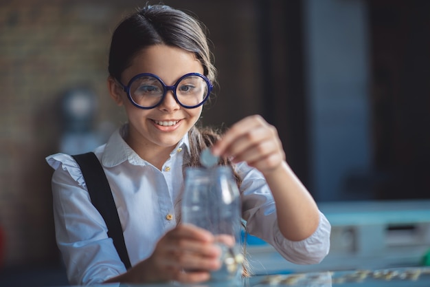 Excité. Fille aux cheveux noirs mettant des pièces dans le bocal et à la recherche d'excitation