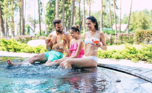Excité heureux et belle jeune famille moderne dans la piscine d'été pendant les vacances à l'hôtel s'amusent et boivent des cocktails.