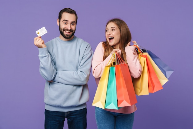 excité heureux couple d'amoureux optimiste isolé sur un mur violet tenant des sacs à provisions et une carte de crédit.