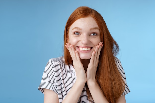 Excité heureux joyeux roux fille au gingembre souriant joyeusement toucher les joues surpris recevoir incroyable cadeau parfait b-day debout étonné triomphant se réjouissant debout fond bleu. Copier l'espace