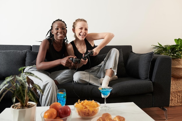 Photo excité jeune couple lesbien diversifié mangeant des collations buvant des cocktails et jouant au jeu vidéo à la maison