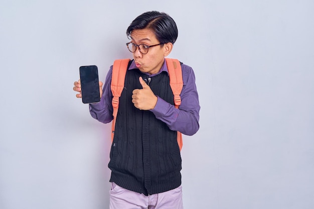 Excité jeune étudiant asiatique en vêtements décontractés avec sac à dos montrant un téléphone portable à écran blanc et un geste du pouce vers le haut isolé sur fond gris Concept de collège universitaire de lycée