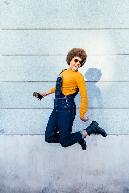 Excité de jeune femme noire sautant avec un smartphone.