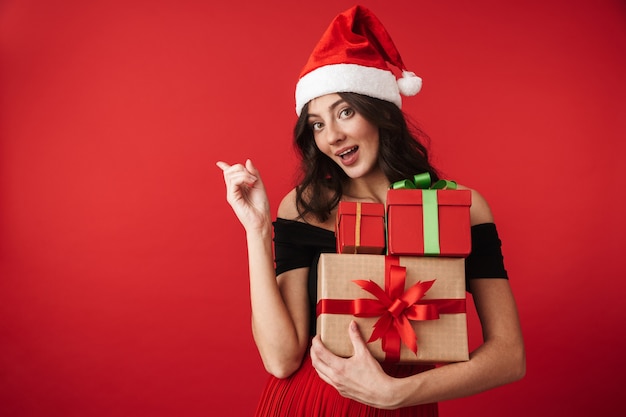 Excité jeune femme portant un chapeau de Noël debout isolé sur rouge, tenant la pile de coffrets cadeaux