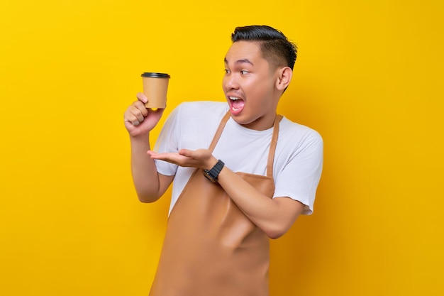 Excité jeune homme asiatique barista barman employé portant un tablier marron travail dans un café montrant une tasse de café ou de thé en papier à portée de main isolé sur fond jaune Démarrage d'une petite entreprise