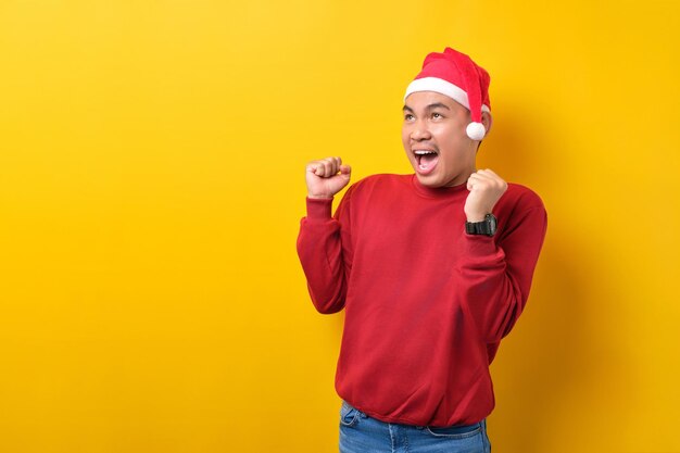 Excité jeune homme asiatique en bonnet de noel levant les mains vers le haut célébrant le succès sur fond de studio jaune célébration vacances de Noël et concept du nouvel an