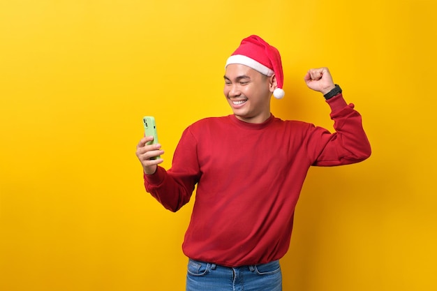 Excité jeune homme asiatique en bonnet de noel utilisant un smartphone et faisant un geste gagnant sur fond de studio jaune célébration vacances de Noël et concept du nouvel an