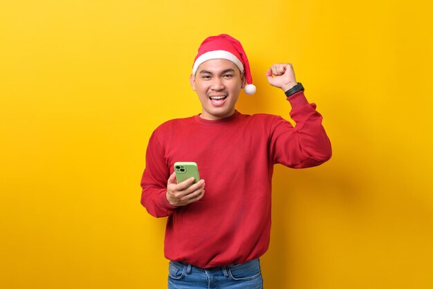 Excité jeune homme asiatique en bonnet de noel utilisant un smartphone et faisant un geste gagnant sur fond de studio jaune célébration vacances de Noël et concept du nouvel an