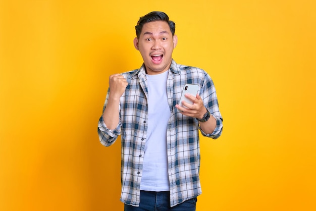 Excité jeune homme asiatique en chemise à carreaux célébrant le succès avec un téléphone portable levant les mains se réjouit grande chance isolé sur fond jaune