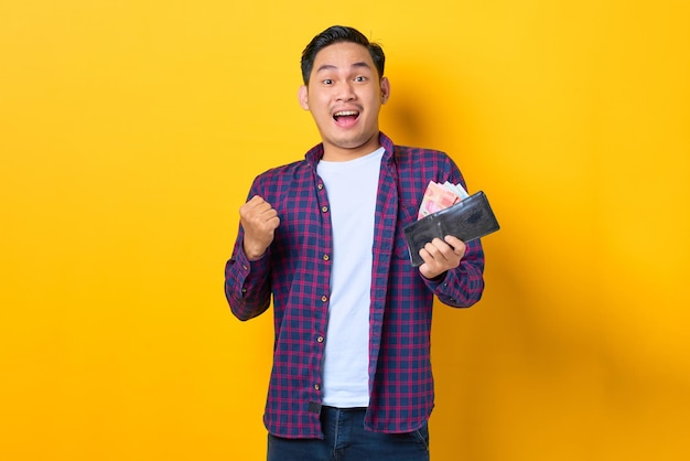 Excité jeune homme asiatique en chemise à carreaux tenant un portefeuille plein de billets de banque isolés sur fond jaune