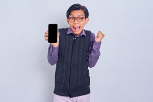 Excité jeune homme asiatique en chemise décontractée et gilet montrant un téléphone portable à écran blanc et faisant un geste gagnant isolé sur fond blanc concept de style de vie des gens