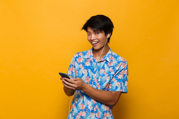 Excité jeune homme asiatique debout isolé sur un espace jaune à l'aide de musique d'écoute de téléphone mobile avec des écouteurs.