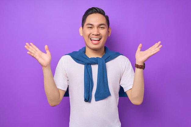Excité jeune homme asiatique portant un t-shirt blanc haussant les épaules avec les mains écartées isolé sur fond violet concept de style de vie des gens