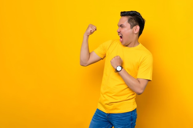 Excité jeune homme asiatique en t-shirt décontracté célébrant le succès avec le poing levé isolé sur fond jaune Concept de style de vie des gens