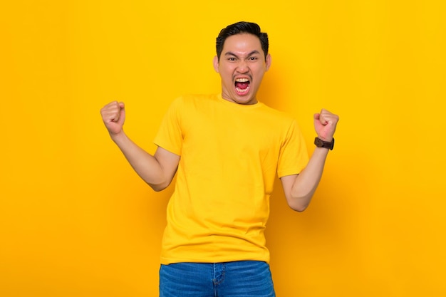 Excité jeune homme asiatique en t-shirt décontracté célébrant le succès avec le poing levé isolé sur fond jaune Concept de style de vie des gens