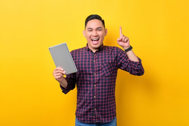 Excité jeune homme asiatique tenant une tablette numérique et levant le doigt vers le haut ayant une bonne idée isolée sur fond jaune