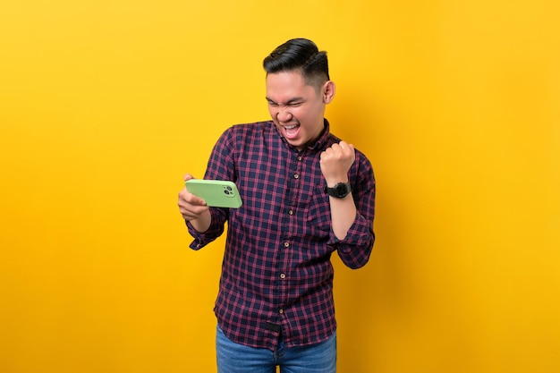 Excité jeune homme asiatique utilisant un smartphone pour jouer à un jeu mobile célébrant le succès isolé sur fond jaune
