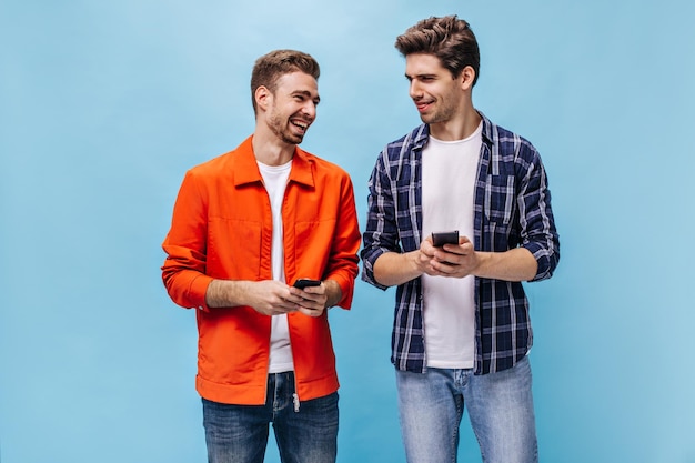 Excité jeune homme barbu en veste orange et son ami en chemise à carreaux tenir le téléphone et rire sur fond bleu