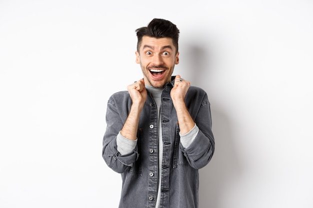 Excité jeune homme sautant du bonheur, souriant étonné et tenant les mains pressées contre la poitrine, debout sur fond blanc.