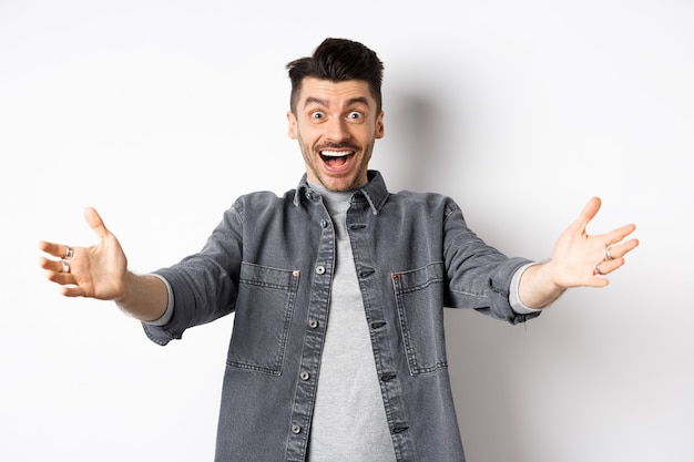 Excité jeune homme tendre les mains pour un câlin, vous câliner, accueillir les invités et sourire, inviter les gens, debout sur fond blanc.