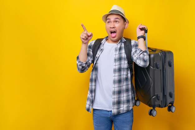 Excité jeune voyageur touriste homme asiatique de 20 ans porte un chapeau de vêtements décontractés avec sac à dos portant une valise pointant le doigt et regardant de côté sur fond jaune Concept de voyage de vol aérien