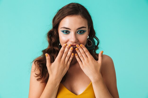 Excité jolie femme portant du maquillage de mode et des boucles d'oreilles souriant et couvrant la bouche avec les mains