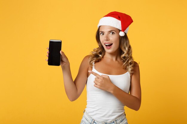 Excité Santa girl in Christmas hat isolé sur fond jaune. Bonne année 2020 concept de vacances de célébration. Maquette de l'espace de copie. Pointant l'index sur le téléphone portable avec un écran vide vide.