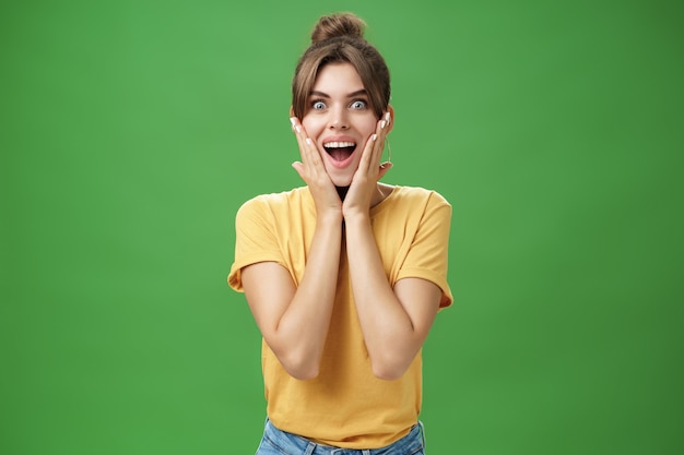 Excité et surpris femme glamour attrayante énergisée en t-shirt jaune touchant les joues d'étonnement souriant largement étonné posant sur le mur vert. Concept d'émotions