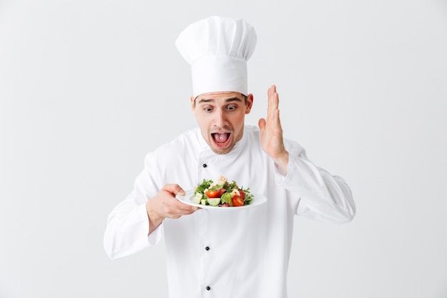 Excited man chef cuisinier en uniforme montrant salade verte fraîche sur une assiette isolée sur mur blanc