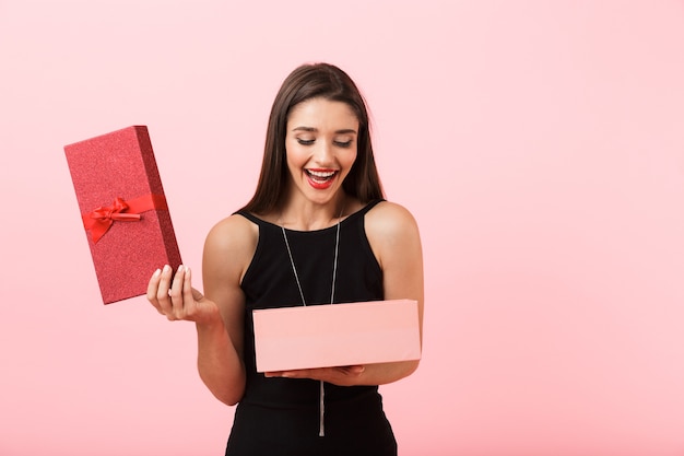 Excited woman wearing black dress holding boîte-cadeau ouvert isolé sur fond rose